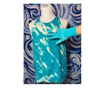 May include: A turquoise blue sleeveless top with a white tie-dye pattern. The top is on a mannequin and has a white flower brooch on the front.