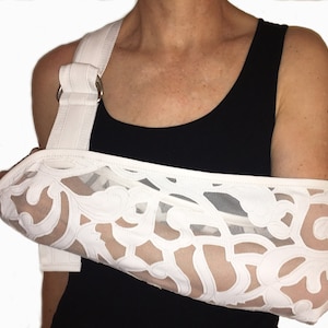 Puede incluir: Funda de brazo blanca con un patrón decorativo de encaje. La funda tiene una correa blanca con una hebilla y dos anillos metálicos.