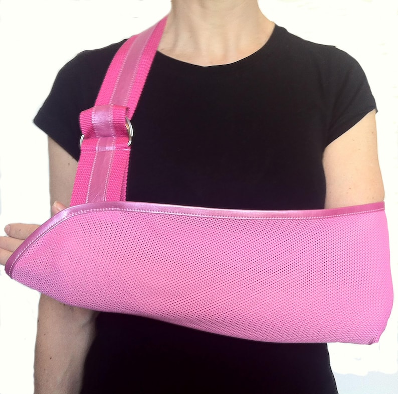 Puede incluir: Un cabestrillo rosa con una correa para el hombro. El cabestrillo est&aacute; hecho de un material de malla y tiene una correa para el hombro acolchada. El cabestrillo est&aacute; dise&ntilde;ado para sujetar un brazo lesionado. La persona de la imagen lleva una camiseta negra.