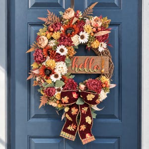 Peut inclure: Une couronne ronde composée de fleurs artificielles aux tons automnaux rouge, orange, jaune et crème, agrémentée de feuillage vert et de baies. Un panneau en bois portant le mot hello est centré, avec un ruban en velours rouge foncé à motifs de feuilles.