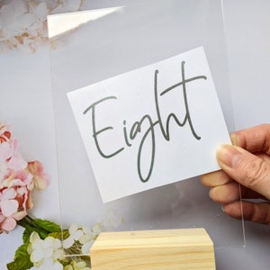 Puede incluir: Porta números de mesa de acrílico transparente con una tarjeta blanca que dice "Eight" en escritura cursiva negra. El soporte está sobre una base de madera.