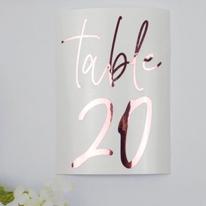 Op de afbeelding: Een witte tafelnummerbord met ros&eacute;gouden metallic letters. Het woord 'table' is in een cursief lettertype boven het nummer '20' geschreven. Het bord is licht gebogen en geplaatst tegen een witte achtergrond met bloemaccenten.