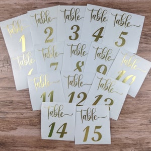 May include: A collection of white table number cards with gold foil lettering. Each card displays the word "Table" in a script font above a number from 1 to 15. The cards are arranged on a wooden surface.