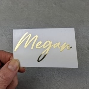 Puede incluir: Pegatina de lámina dorada con el nombre Megan escrito en cursiva.