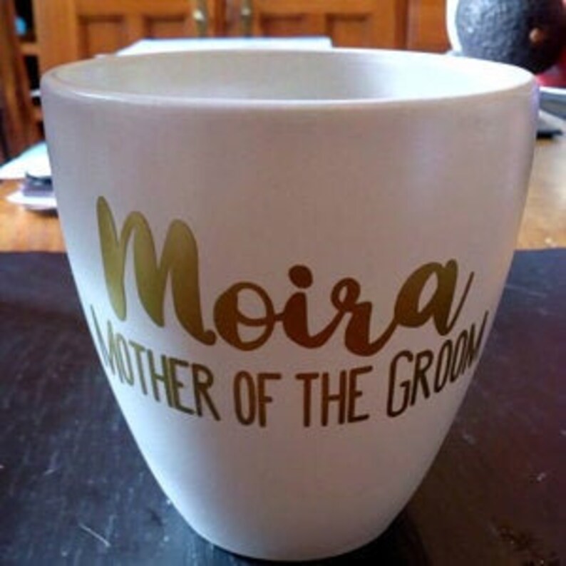 May include: White ceramic mug with gold lettering that reads "Moira Mother of the Groom".