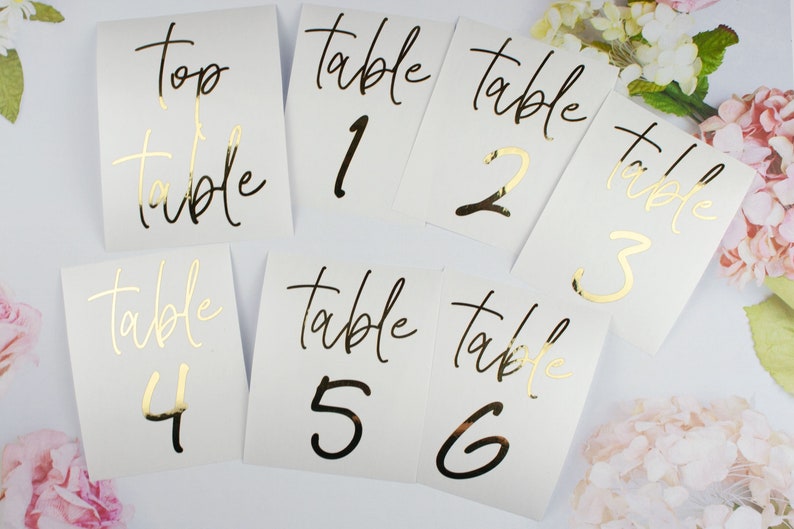 Op de afbeelding: Witte tafelnummerkaarten met gouden en zwarte letters. De kaarten zijn genummerd van 1 tot en met 6, met het woord "tafel" boven elk nummer. E&eacute;n kaart leest "top table". De kaarten zijn gerangschikt op een wit oppervlak met bloemaccenten.