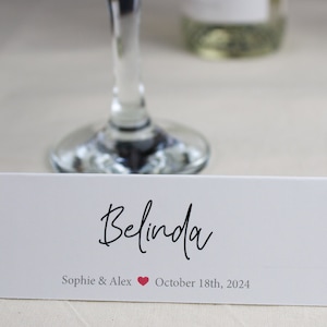 May include: White place card with black cursive text "Belinda" and the text "Sophie & Alex" with a red heart symbol and "October 18th, 2024".