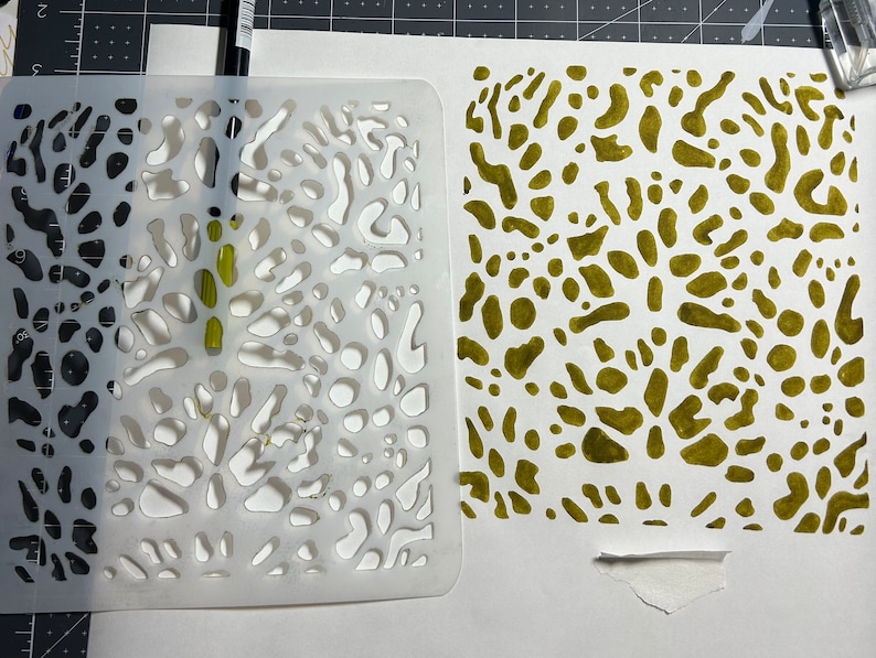 May include: A white plastic stencil with a pattern of irregular shapes, and a green painted sheet of paper with the same pattern.