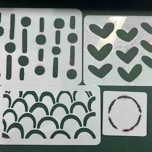 May include: Four white plastic stencils with different designs. The first stencil has a pattern of circles and lines. The second stencil has a pattern of hearts. The third stencil has a pattern of curved lines. The fourth stencil has a circle with a textured edge.