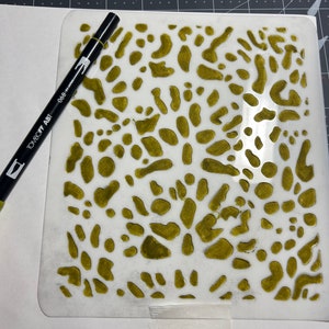 May include: A white stencil with a green paint design of abstract shapes. The stencil is on a white surface with a black marker pen.