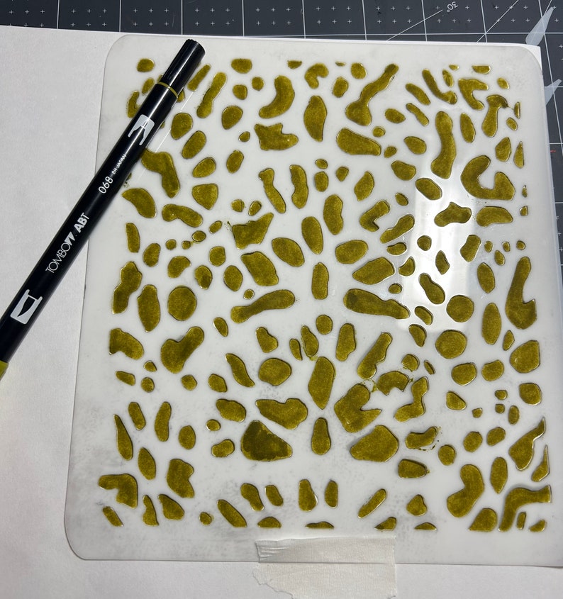 May include: A white stencil with a green paint design of abstract shapes. The stencil is on a white surface with a black marker pen.