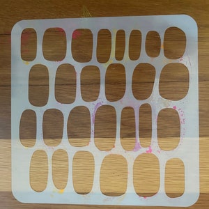 Puede incluir: Plantilla de plástico blanco con 20 recortes ovalados dispuestos en una cuadrícula. La plantilla está sobre una superficie de madera con salpicaduras de pintura.