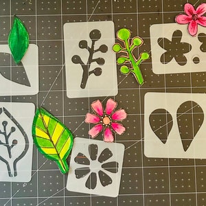 May include: A collection of stencils for crafting, featuring various floral and leaf designs. The stencils are made of white plastic and are arranged on a black grid surface. The designs include flowers, leaves, and branches.