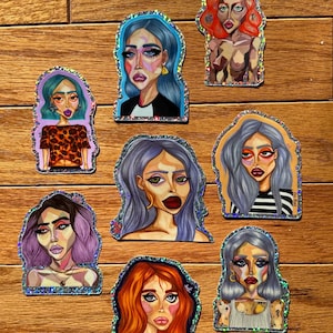 May include: A collection of colorful stickers featuring stylized portraits of women with bold makeup and various hair colors, including blue, red, and purple. Each sticker has a glittery border and is set against a wooden background.