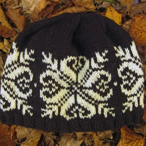 Può includere: Un berretto lavorato a maglia marrone scuro con un motivo a fiocco di neve bianco e giallo chiaro. Il cappello è realizzato in un materiale morbido e strutturato ed è adatto al clima freddo. Il berretto è appoggiato su foglie autunnali.