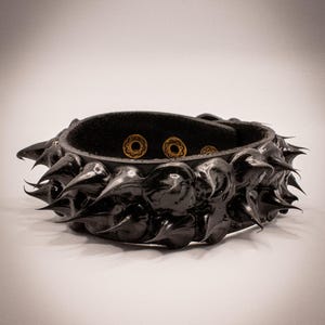 May include: Black leather bracelet with large, glossy, pointed spikes. The bracelet has a rounded shape and features three gold-colored decorative rivets. The spikes are evenly spaced around the exterior of the bracelet.