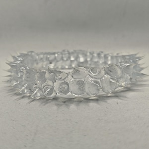 May include: A clear, transparent ring with a textured surface. The ring features a series of rounded, bubble-like shapes and pointed spikes around the outer edge. The ring is made of a clear material and is set against a neutral background.