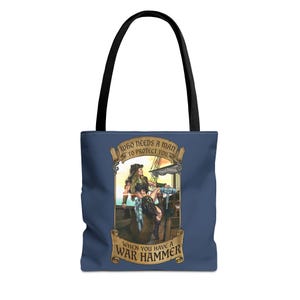 May include: A navy blue tote bag with black handles featuring a graphic illustration of a warrior sitting on a barrel on a ship deck, holding a war hammer. The text reads: WHO NEEDS A MAN TO PROTECT YOU WHEN YOU HAVE A WAR HAMMER.
