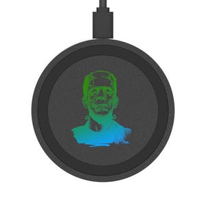 Op de afbeelding: Een zwarte draadloze oplader met een groen-blauwe illustratie van Frankenstein's monster. De oplader heeft een cirkelvormig ontwerp met een zwarte rand en een USB-kabel.
