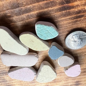 May include: A collection of small, irregularly shaped, pastel-colored stones, including shades of pink, blue, green, and cream, are displayed on a wooden surface. A U.S. quarter coin is included for size comparison. The stones have a weathered appearance.