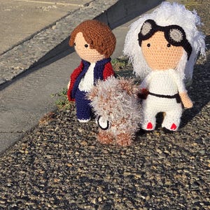 May include: Three handmade crochet dolls. One doll has brown hair, a blue jacket with red accents, and a white shirt. Another doll has white hair, goggles, and a white jumpsuit. A small, brown, furry creature is also present.