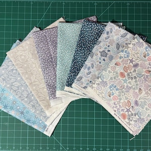 May include: A selection of fabric squares featuring marine-themed designs, such as starfish and shells, in blue, grey, and beige hues. The fabric is displayed on a green cutting mat.