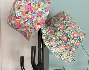 May include: Two patterned sun hats. One hat features a colorful village scene with houses, trees, and flowers. The other hat has a floral design with pink flowers, green leaves, and yellow accents. Both hats are made of fabric and are hanging on a black coat rack.