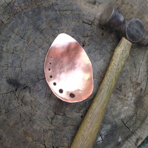 May include: A copper metal piece with a hammered texture and several drilled holes, next to a ball-peen hammer with a wooden handle. The metal piece has an organic, teardrop shape. The background is a weathered wooden surface.