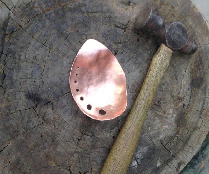 May include: A copper metal piece with a hammered texture and several drilled holes, next to a ball-peen hammer with a wooden handle. The metal piece has an organic, teardrop shape. The background is a weathered wooden surface.