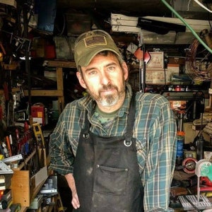 May include: A man wearing a green camouflage hat with an American flag patch, a plaid shirt, and a black apron. The background shows a cluttered workshop with tools and equipment, suggesting a craft or repair setting.