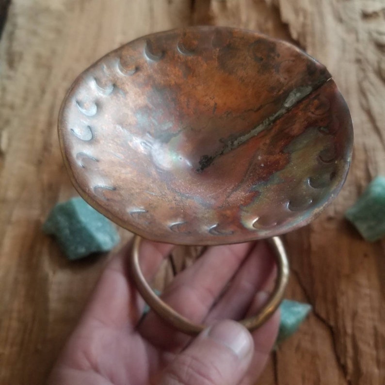 May include: A copper bowl with a hammered texture and a brass ring handle. The bowl has a patina finish and is slightly misshapen.