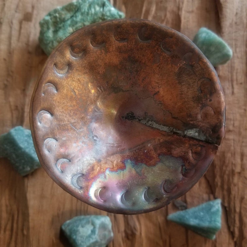 May include: A copper bowl with a hammered finish and a crescent moon design. The bowl has a patina finish and is slightly dented.