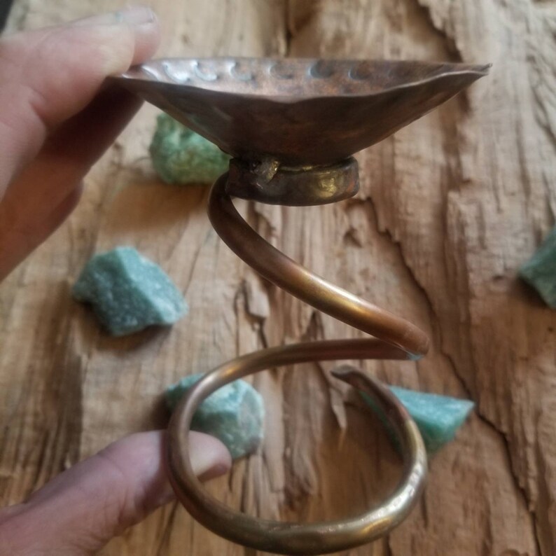 May include: A copper candle holder with a spiral base and a hammered copper dish on top. The holder is on a wooden surface with green stones.