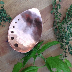 May include: A copper-colored, teardrop-shaped serving dish with several circular holes. Fresh herbs, including thyme and basil, are arranged around the dish on a wooden surface. The dish is likely used for food preparation or serving.