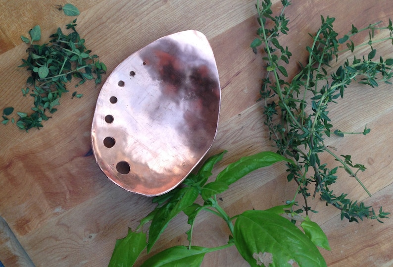 May include: A copper-colored, teardrop-shaped serving dish with several circular holes. Fresh herbs, including thyme and basil, are arranged around the dish on a wooden surface. The dish is likely used for food preparation or serving.