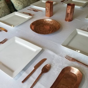 May include: A table setting with white square plates, copper-colored cutlery, and hammered copper serving pieces. The table is set on a white tablecloth. Two copper candle holders are also present.