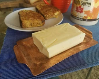 May include: A stick of butter on a copper butter dish, with two pieces of French toast on a white plate in the background.