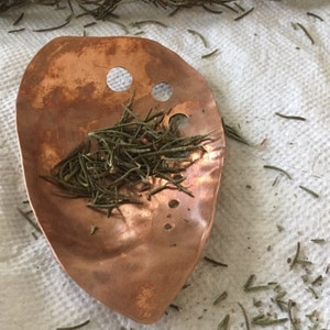 May include: A copper-colored, leaf-shaped scoop with two circular holes near the top, filled with dried rosemary. The scoop rests on a white, textured surface, with scattered rosemary sprigs around it. The scoop has a hammered texture.