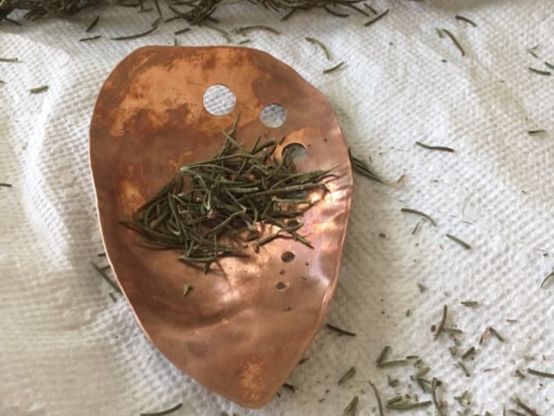 May include: A copper-colored, leaf-shaped scoop with two circular holes near the top, filled with dried rosemary. The scoop rests on a white, textured surface, with scattered rosemary sprigs around it. The scoop has a hammered texture.