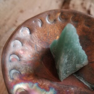 May include: A green crystal shaped like a pyramid sits on a copper dish with a crescent moon design.