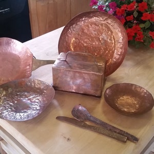 May include: A collection of handcrafted copper kitchenware, including a pan, bowls, a plate, a box, and utensils. The items have a hammered texture and a warm, reddish-brown color, suggesting a rustic or vintage aesthetic. The arrangement is on a light-colored wooden surface.