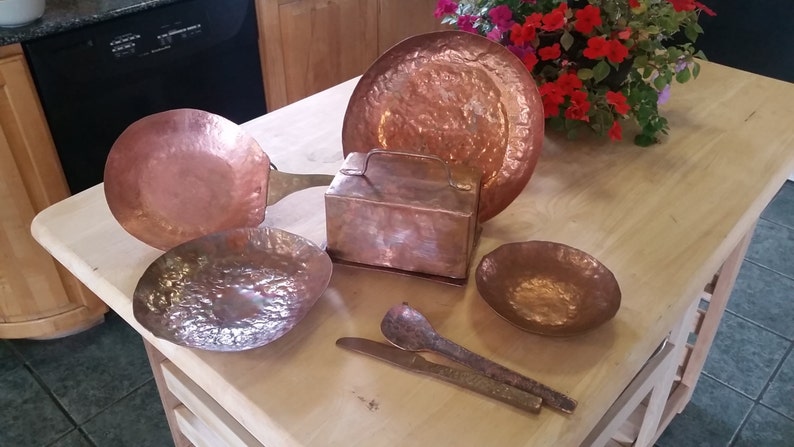 May include: A collection of handcrafted copper kitchenware, including a pan, bowls, a plate, a box, and utensils. The items have a hammered texture and a warm, reddish-brown color, suggesting a rustic or vintage aesthetic. The arrangement is on a light-colored wooden surface.