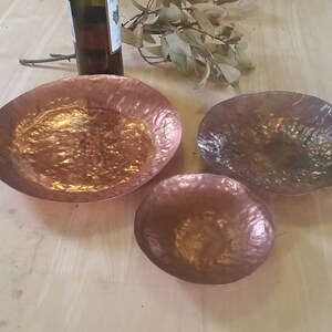 May include: Three copper bowls of varying sizes, with hammered textures, on a wooden surface.