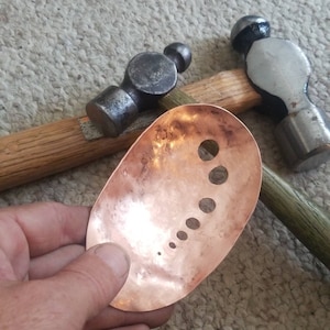 May include: A copper metal piece with several circular cutouts is held in a hand. Two ball-peen hammers with wooden handles are in the background. The metal piece has a polished, reflective surface.