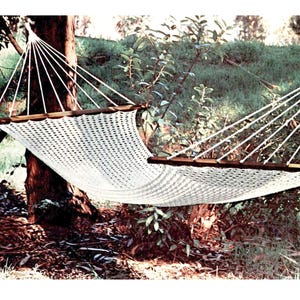 May include: A white woven hammock suspended between two trees in a natural outdoor setting. The hammock has wooden support bars and is made of a durable, open-weave material. The scene is surrounded by lush greenery.