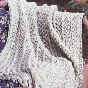 Pode incluir: Uma manta de crochê creme com um padrão de chevron e detalhes de franjas. A manta está sobre uma estrutura de cama rosa com uma almofada estampada floral visível por baixo. A textura e o design da manta são proeminentes.