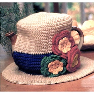 Puede incluir: Un cubreteiera de ganchillo con diseño de sombrero. El cubreteiera es beige con una banda azul y adornado con tres flores de ganchillo rojas, verdes y amarillas. El texto "digital pattern shop" es visible en el ala.