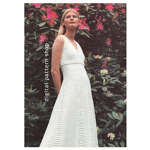 May include: A white crochet dress with a V-neckline and a fitted bodice. The dress has a flared skirt and is decorated with a crocheted trim around the neckline and waist. The dress is photographed on a model standing in front of a bush of pink flowers.