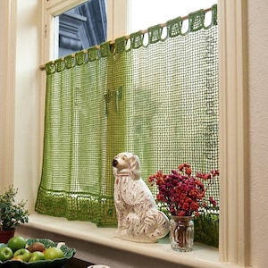 May include: A green crochet cafe curtain with a grid pattern and loop top, hanging on a wooden rod. On the windowsill sits a white ceramic dog figurine, a clear glass vase with dried pink flowers, and a bowl filled with green apples.