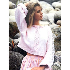 May include: A light pink, cropped sweater with long sleeves. The sweater features a decorative cable knit design down the center. The model is wearing a pink and orange striped skirt. The background is a rocky beach.